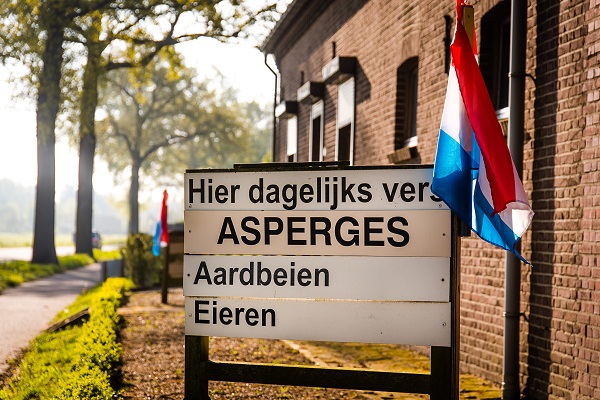 Bord met de tekst ‘Hier dagelĳks vers asperges, aardbeien, kersen’ bij een boerderij met Limburgse vlag, omgeven door bomen en landelijke sfeer.