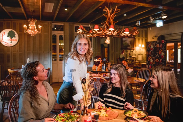 Een serveerster brengt eten naar een groep van drie lachende vrouwen bij Boscafé de Busjop in Heyhuysen met houten interieur en kroonluchters.