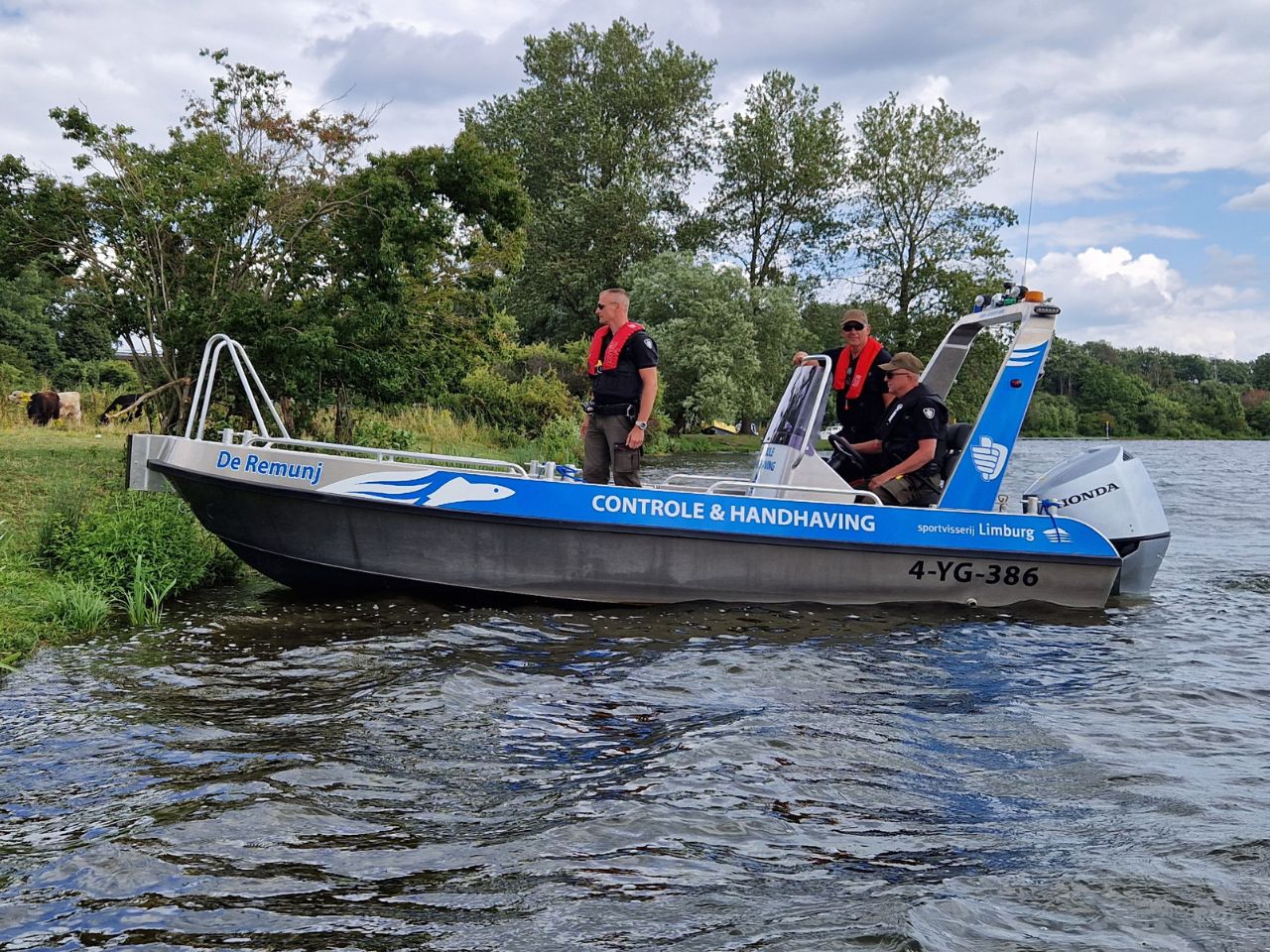Control and enforcement boat “De Remunj” of Sportvisserij Limburg sails along the shore of a pond. Three enforcers in black uniforms and with life jackets are on board. In the background, cattle are grazing in a green nature reserve. The boat has a blue print with the text “Controle & Handhaving” and the registration number 4-YG-386.