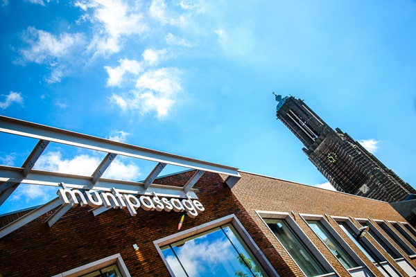 Eingang zum Einkaufszentrum Muntpassage in Weert, mit dem Turm der Sint-Martinus-Kirche im Hintergrund vor einem blauen Himmel mit Wolken