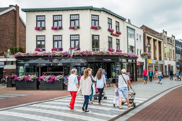Im Zentrum von Weert überqueren die Einkäufer die Straße, vorbei an einer Blumenterrasse und mehreren Geschäften im Hintergrund.