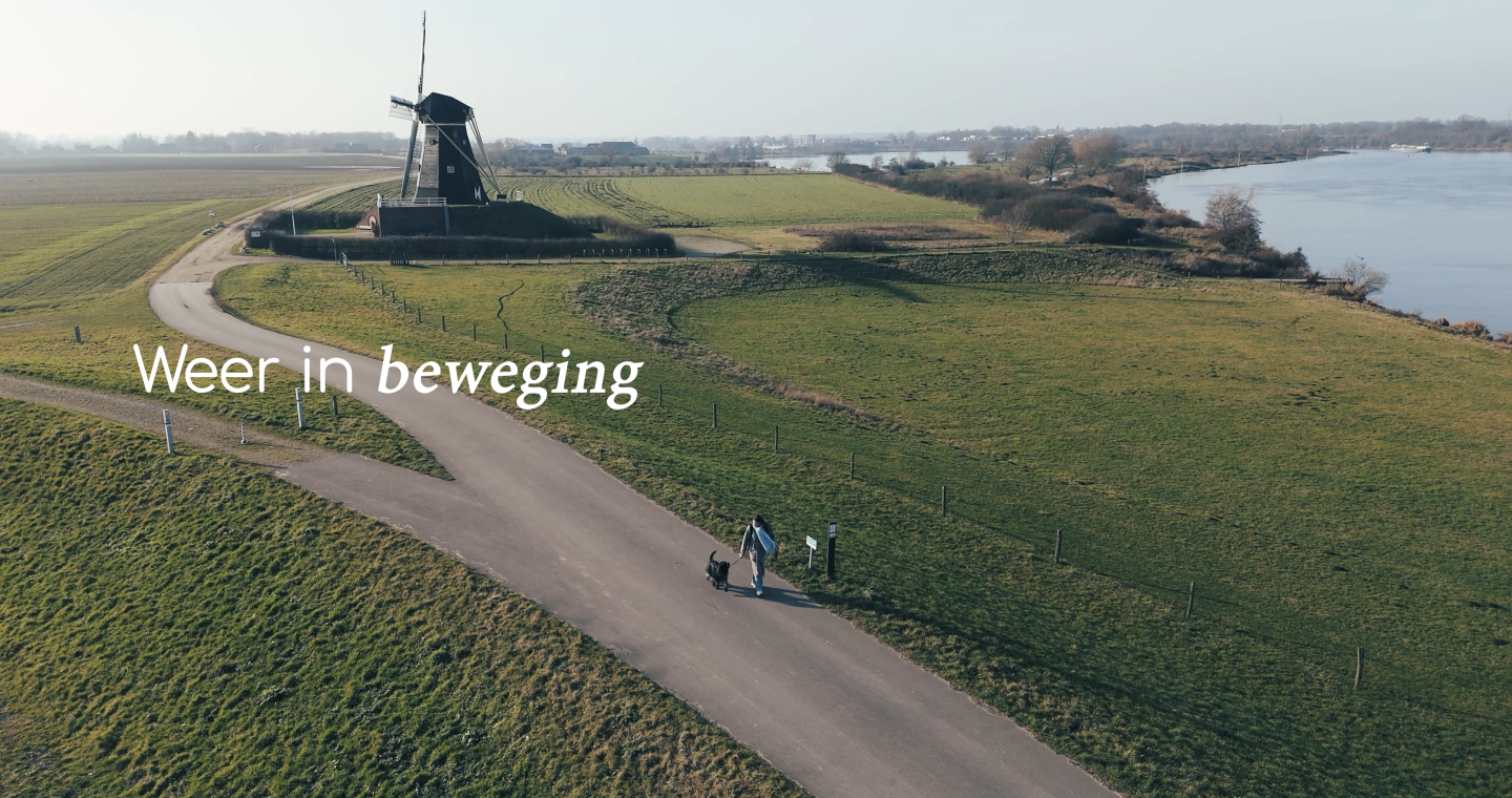 Luchtfoto van een wandelaar met hond op een dijkweg langs de Maas, met een historische windmolen en uitgestrekte velden in Midden-Limburg, met de tekst “Weer in beweging”.
