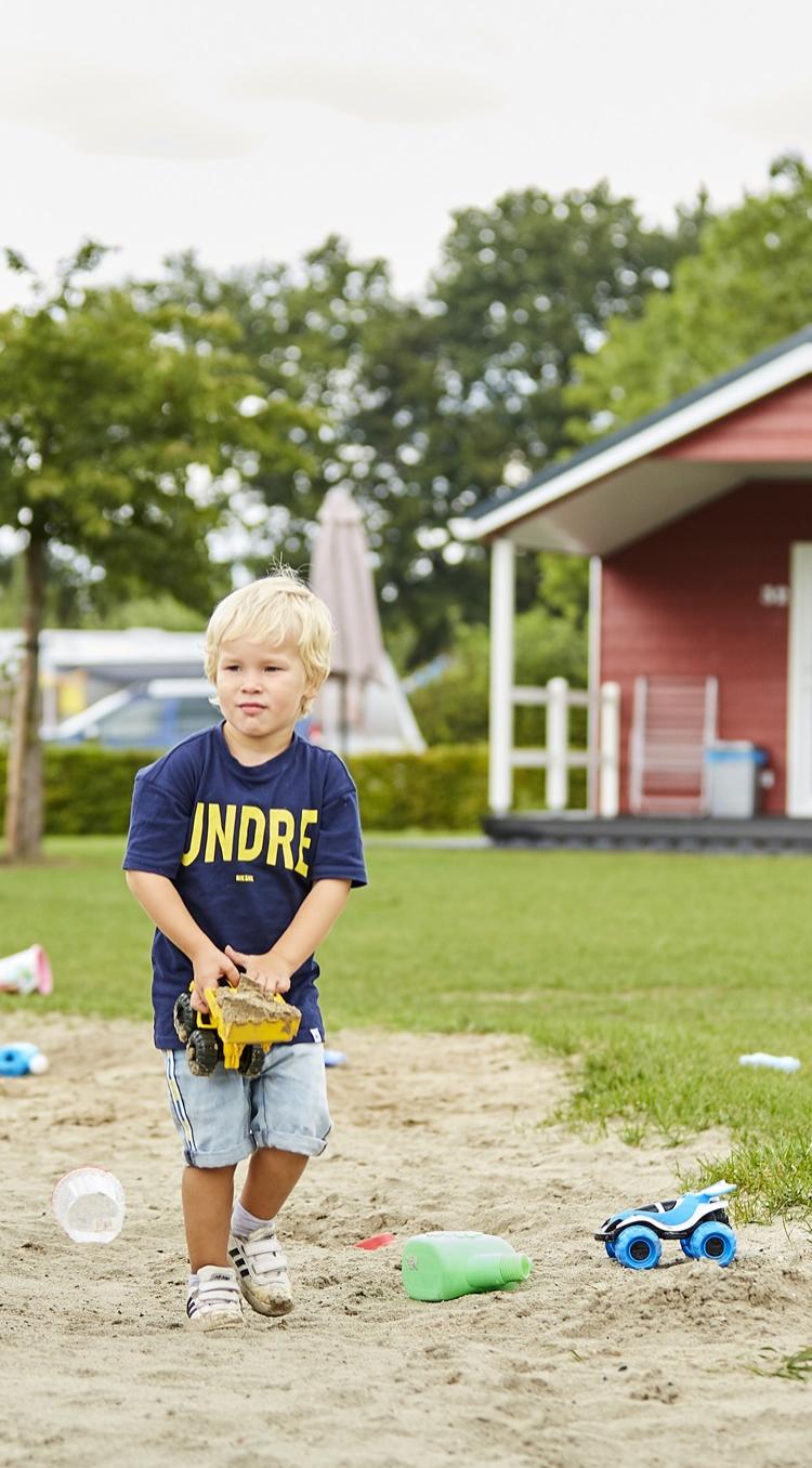 Jong kind speelt met een speelgoedgraafmachine in het zand op Recreatiepark de Leistert, met een vakantiehuisje op de achtergrond.