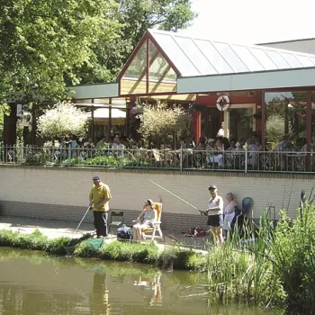 Mensen vissen langs de waterkant bij Forellenvijver Heioord in Neeritter, met op de achtergrond een terras vol bezoekers.
